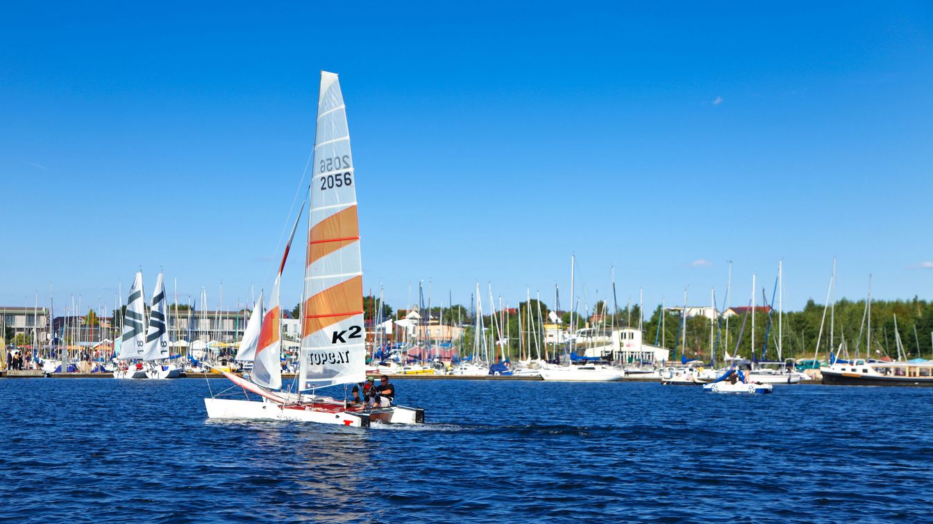 Ein Segelboot auf dem strahlend blauem Wasser des Cospudener Sees im Leipziger Neuseenland, im Hintergrund unzählige weitere Segelboote am Hafen